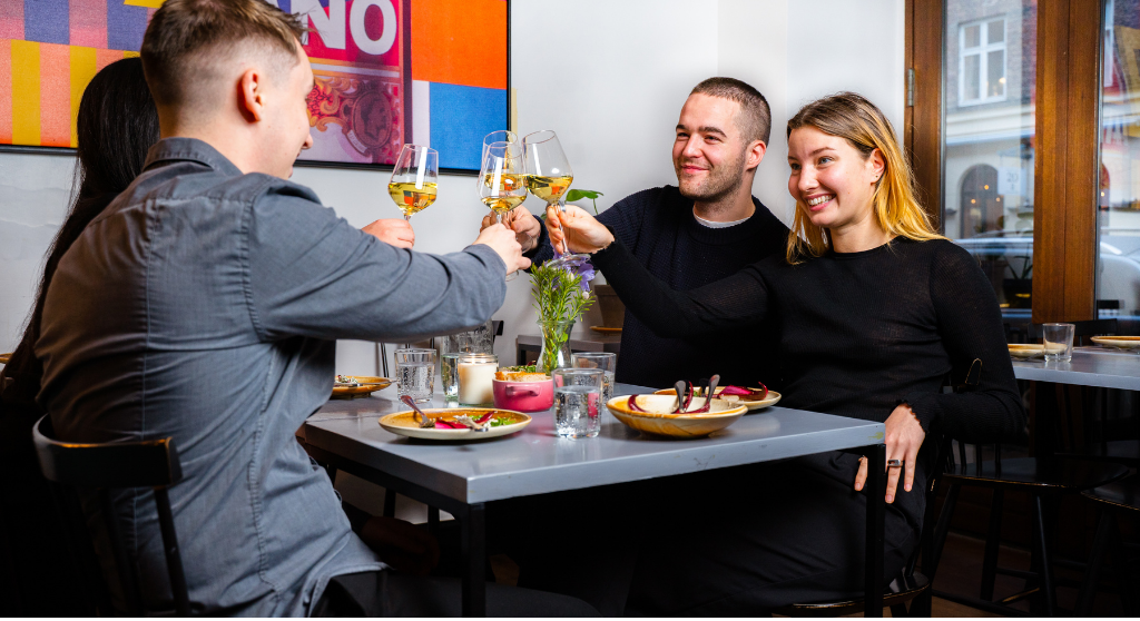 Four smiling people. two women and two men, clinking white wine glasses at a restaurant table. A colourful poster hangs on the wall to the left.