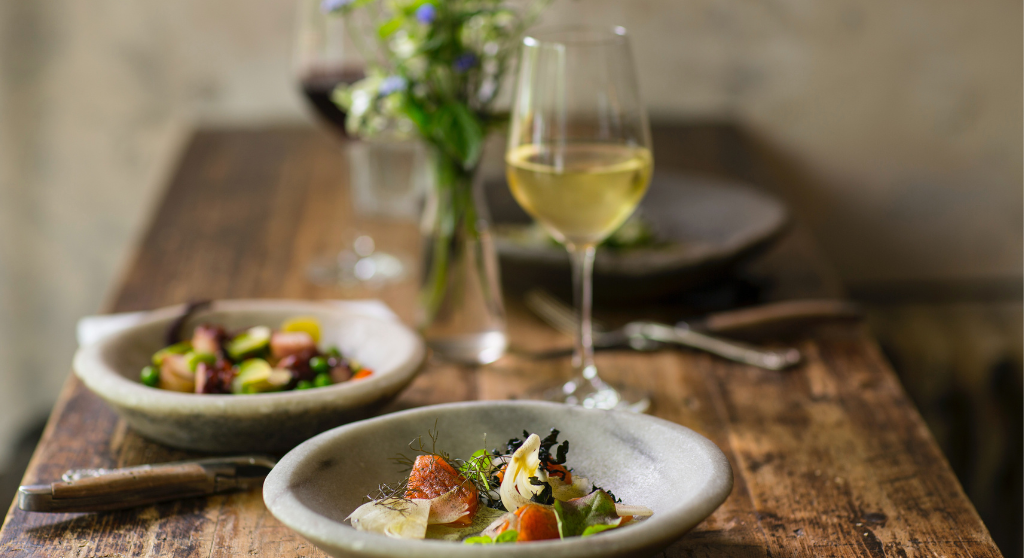 Two plates of food, and two glasses of wine on a wooden table decorated with flowers in a small vase.
