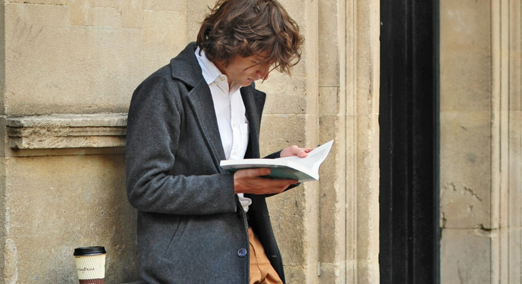 A man leaning against a wall while reading a book. There's a cup of takeaway coffee next to him. 