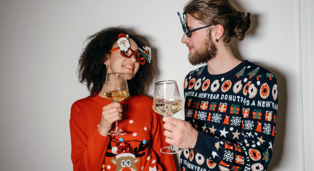 A man and a woman dressed in Christmas sweaters. They are a holding wine glasses. 
