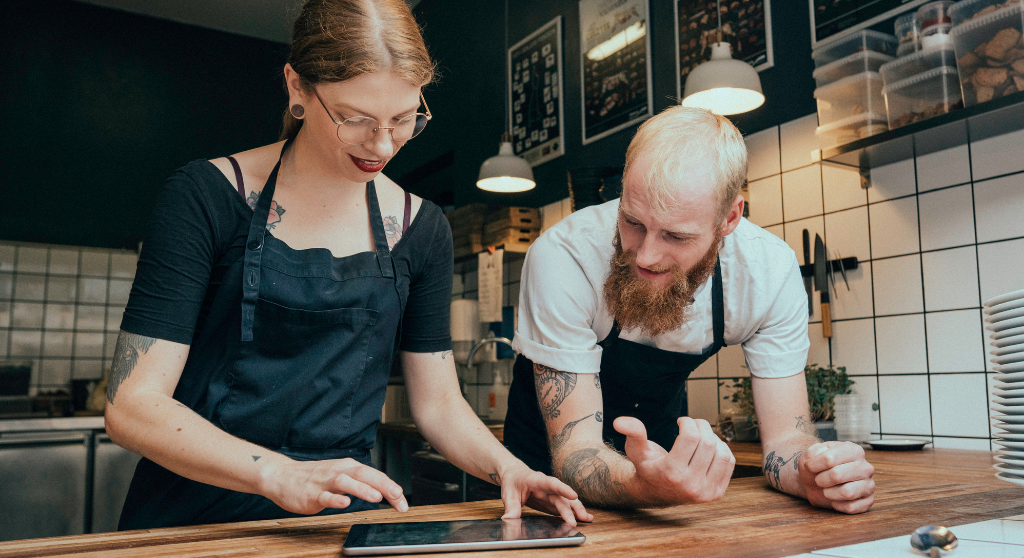 A man and a woman looking at a tablet. They're in a restaurant kitchen and wearing black aprons.