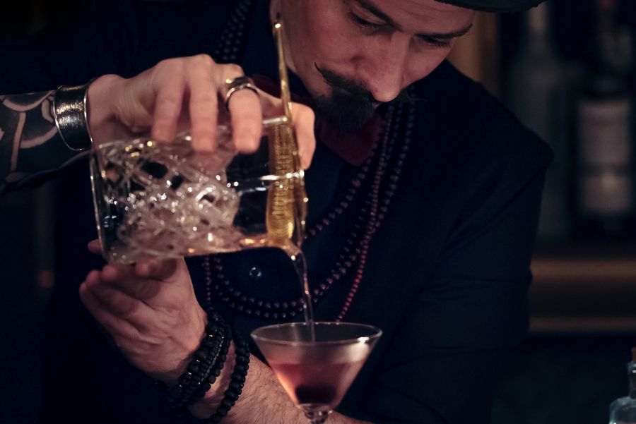 A bartender dressed in black pouring a Halloween cocktail into a glass