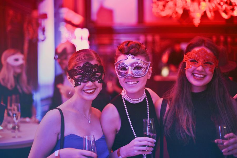Three smiling women wearing carnival masks and drinking champagne. 