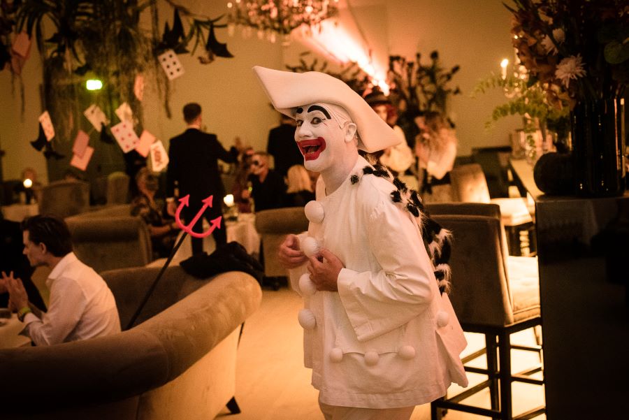Halloween in Nimb Bar, 2021. The pantomime clown, Pierrot, is standing in the bar, with a broad red smile. To the left of him is a guest in an arm chair. The guest has a plastic trident. Playing cards are hanging from the ceiling. 