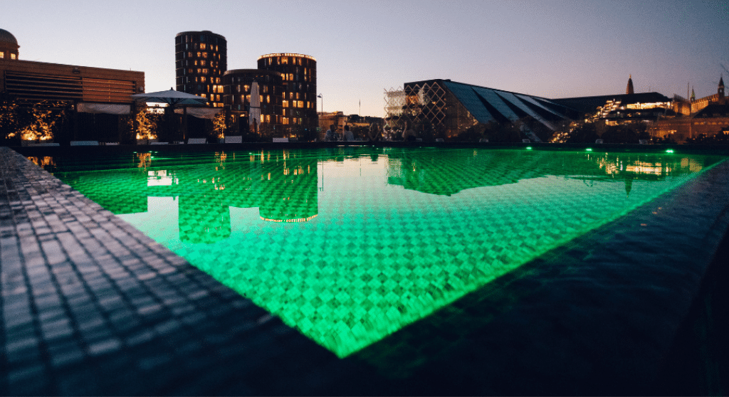 A pool at hotel Nimb's roof-top terrace. The water is illuminated and looks green. Copenhagen's skyline is reflected in the surface. A few people are sitting at tables near the pool.