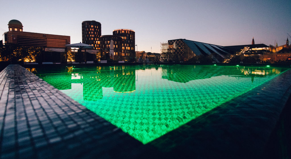 A pool at hotel Nimb's roof-top terrace. The water is illuminated and looks green. Copenhagen's skyline is reflected in the surface. A few people are sitting at tables near the pool