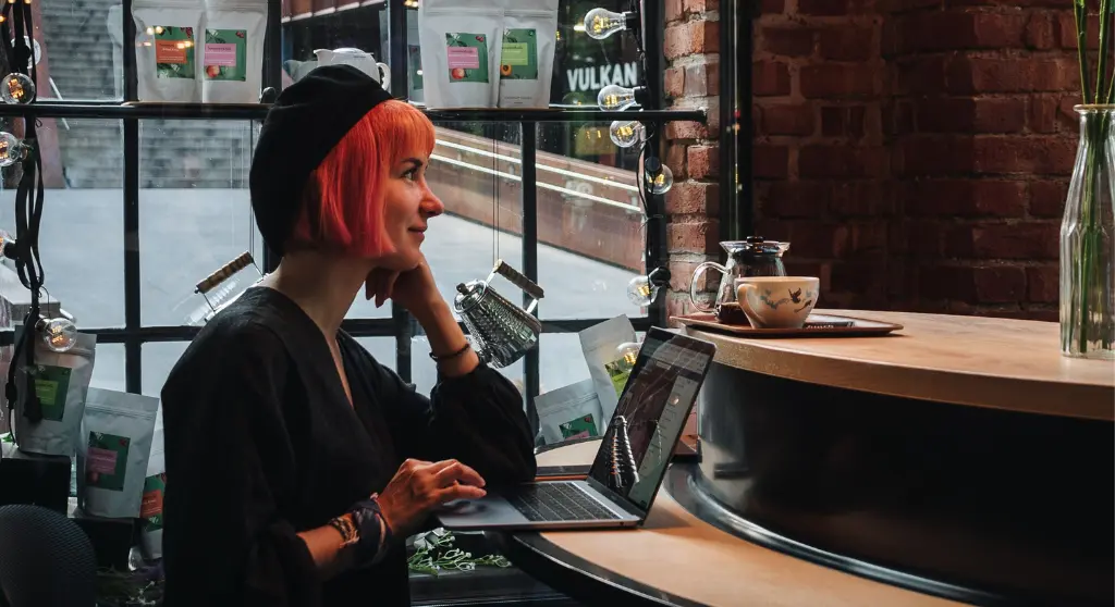 A redhaired woman with a black beret writing on a laptop in a restaurant