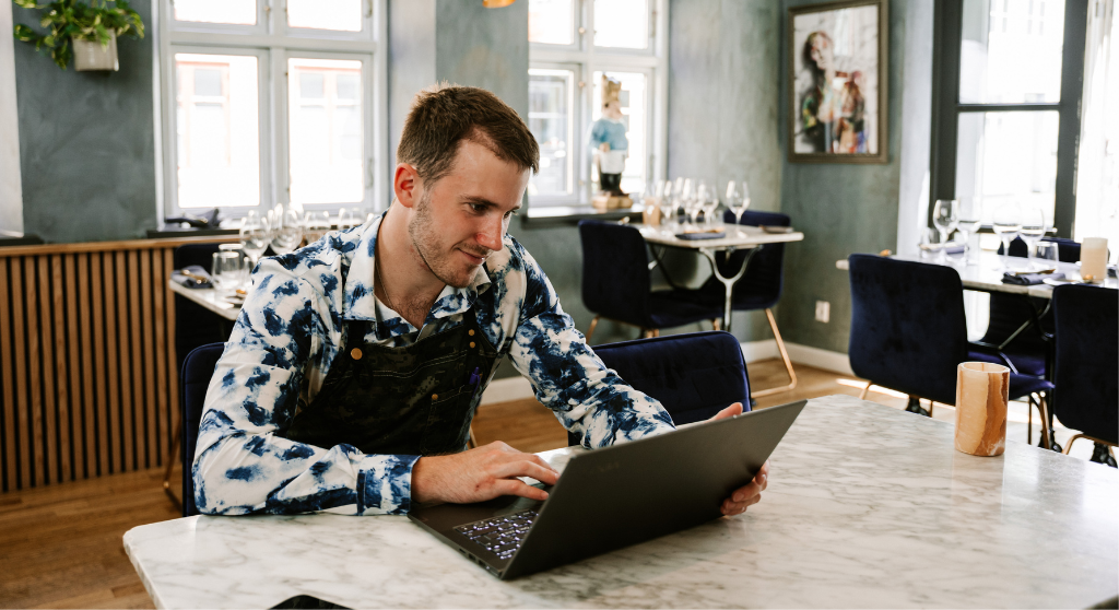 A man using a laptop in a restauant. He's wearing an apron. Behind him are three tables set with wine glasses and cutlery.