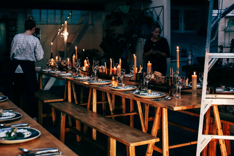 A long wooden table set with plates, glasses, sliced bread on trays and many lit candles. 