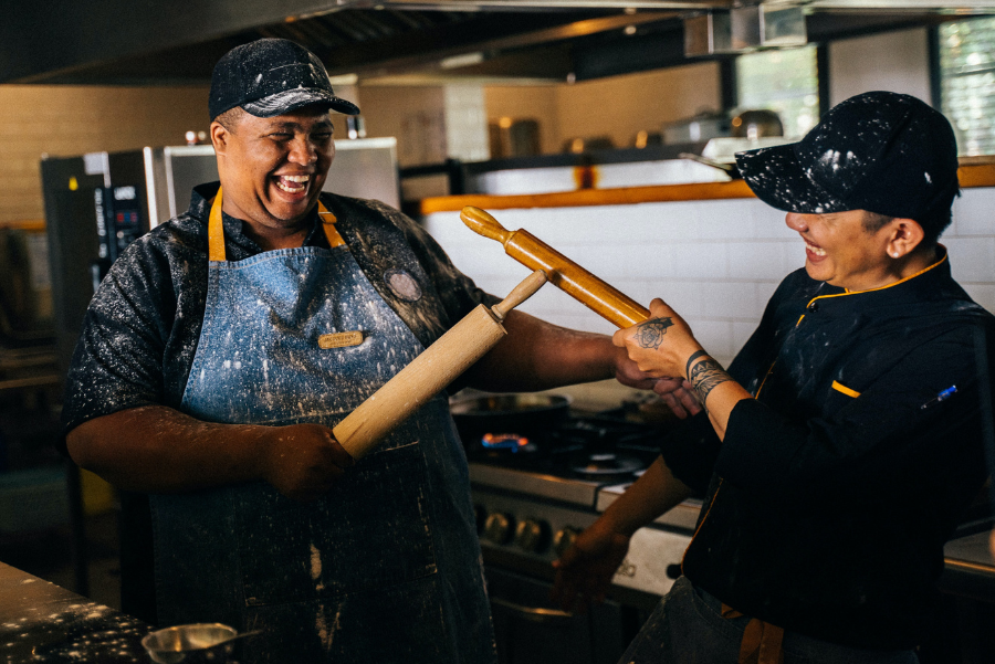 Two laughing cooks pointing rolling pins at each other in the kitchen. Both have flour sprinkled on their dark blue uniforms. 