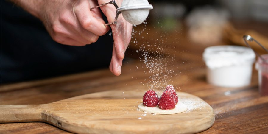 Hands using a tea sieve to sprinkle powdered sugar over three raspberries on a circle of pastry dough. 